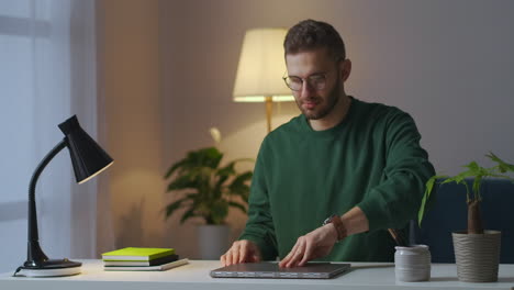 young-man-with-glasses-is-using-laptop-at-evening-at-home-opening-notebook-and-switching-on-table-lamp-medium-portrait-of-guy-with-glasses
